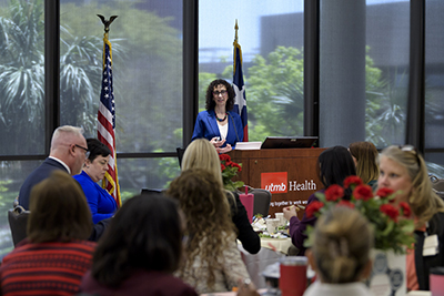 Dr. Suzie Bergman stands at the podium speaking to a room of people seated at tables.