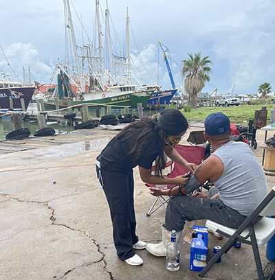 A student in black scrubs places a blood pressure cuff on a man outdoors next to a boat
